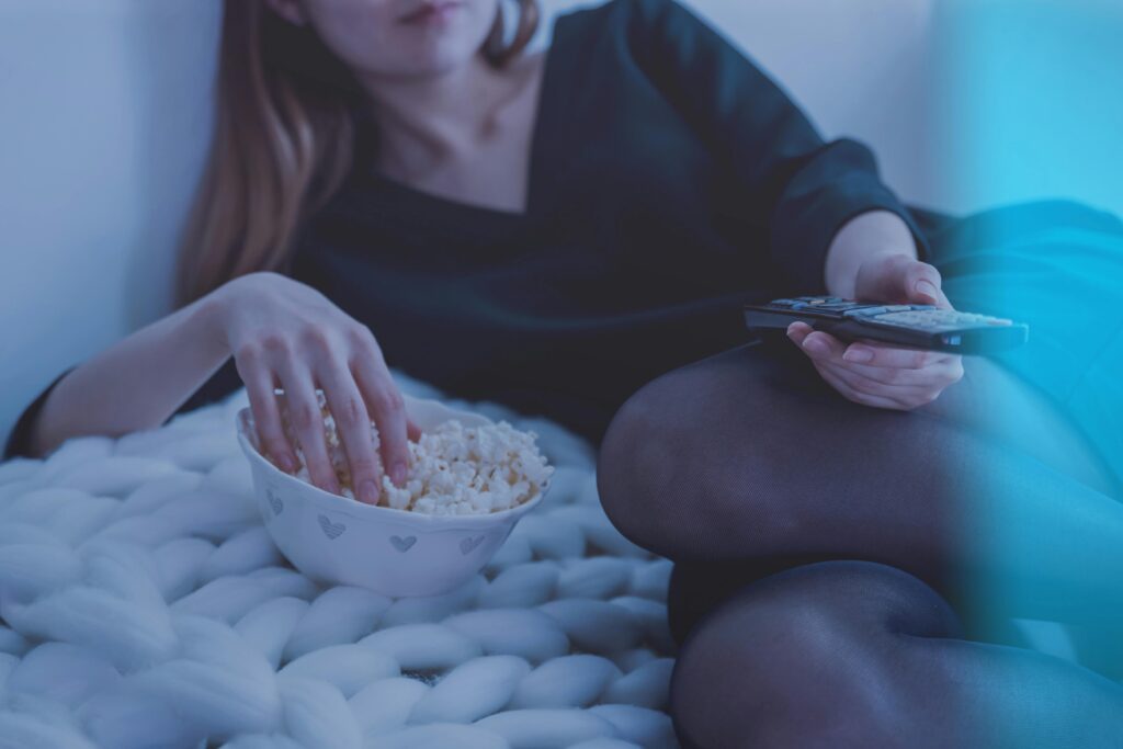 Lady eating with hand in bowl of popcorn, other hand holding remote control, while laying down