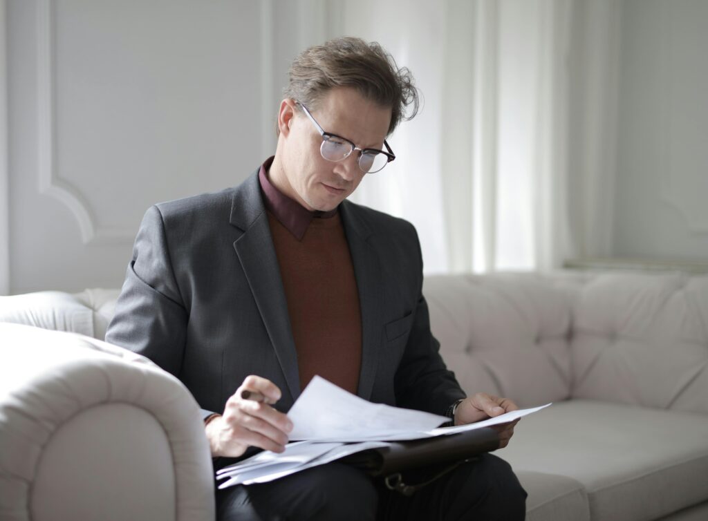 Man sitting on a couch with papers for a durable power of attorney on this lap, pen in hand. Man is wearing eyeglasses and a suit. 
