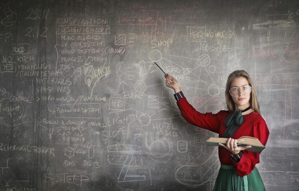 a teaching pointing to a chalkboard. She is holding a book in the other hand. On the chalkboard is scribbles and drawings