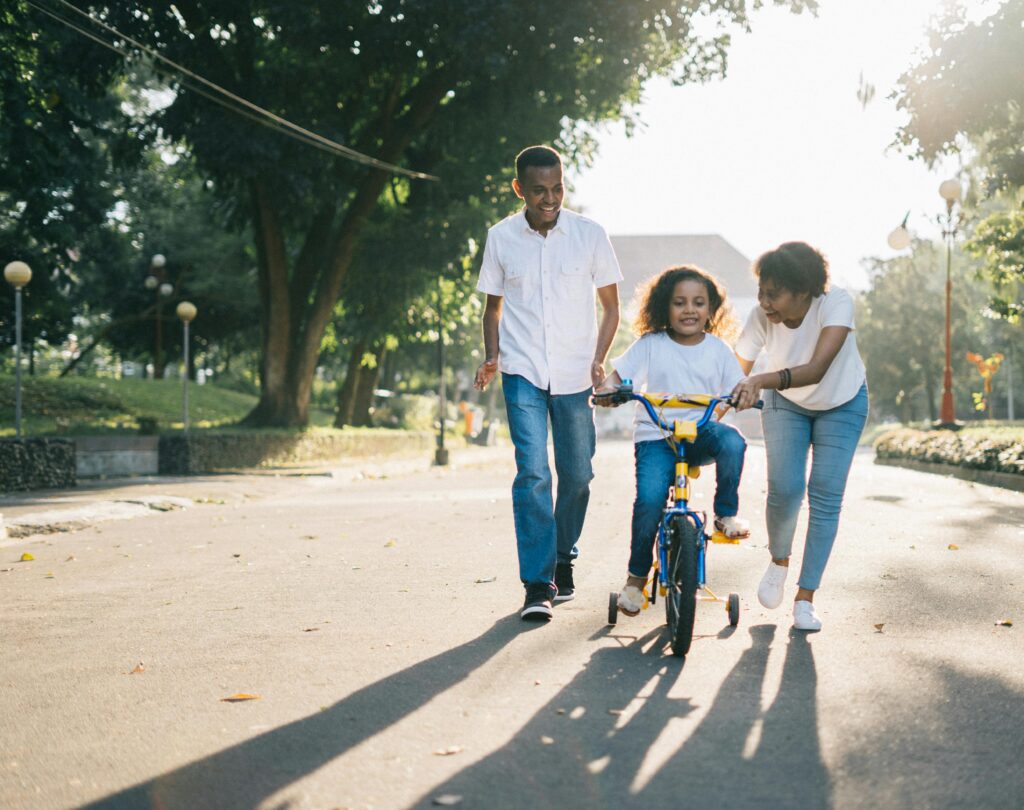 man and women teaching small girl how to ride a bike. woman is holding bike while girl pedals. 