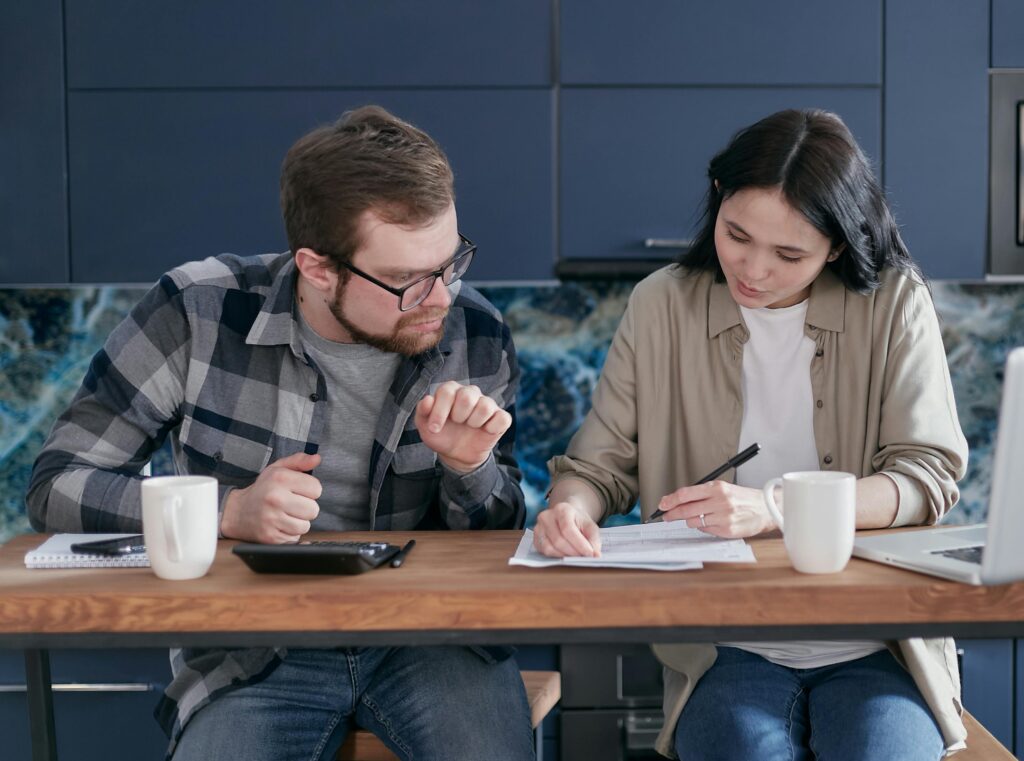 lady and man sitting at a table looking at a paper. Lady is holding a pen. each has a coffee cup next to them. 