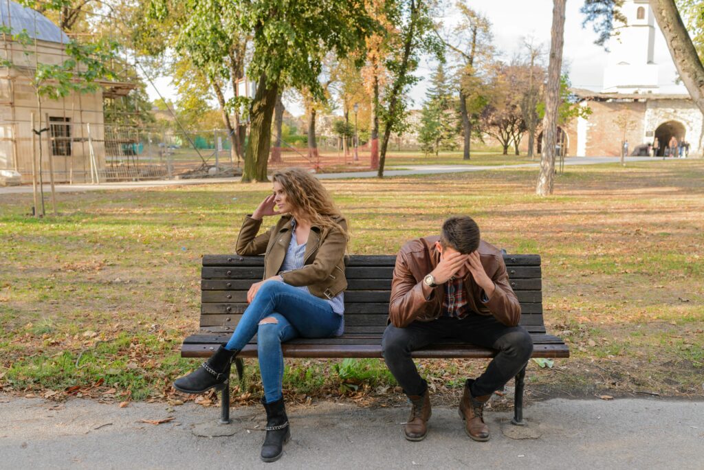 Man and lady sitting on park bench. Man has hands on face, woman is turned the other direction.