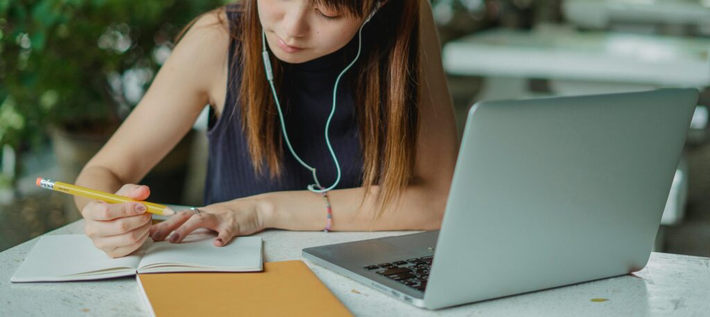 girl writing in a notebook with laptop open on a table. 