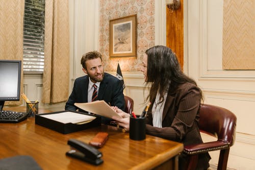 Man and woman sitting at a desk and woman is holding papers. Suitcase is on the desk open with more papers. 